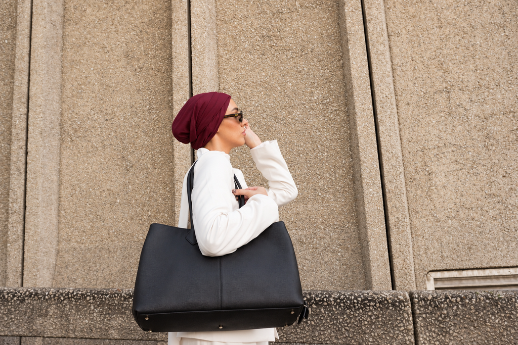 A woman in a white outfit and burgundy headscarf carries a large black tote bag against a textured beige stone wall background.