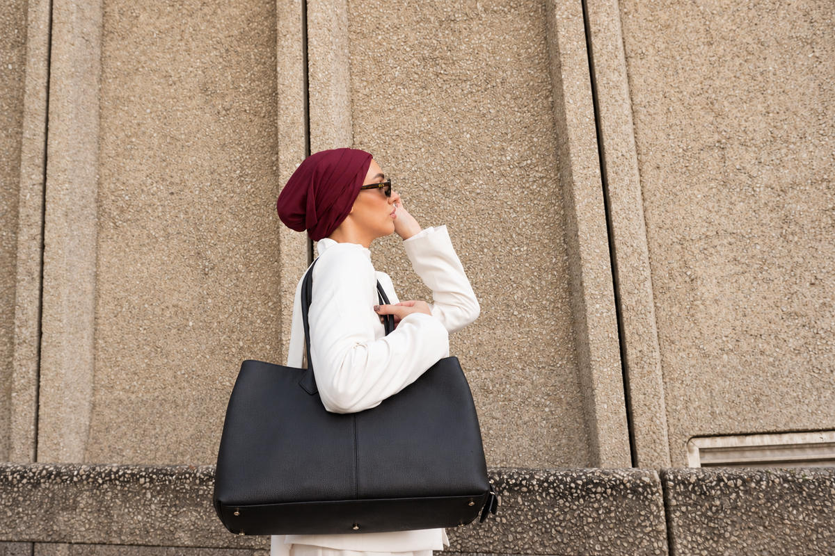A woman in a white outfit and burgundy headscarf carries a large black tote bag against a textured beige stone wall background.