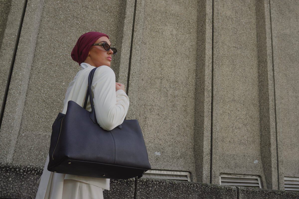 A woman wearing a burgundy headscarf, white outfit, and black sunglasses carries a large black leather tote bag against a textured concrete wall.